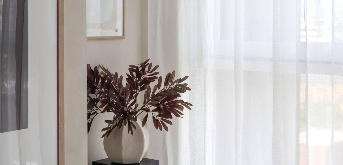 Corner of a living room showing a large beige vase with some dried leaves in a black plant stand. Neutral colours on the walls with matching artwork hanging. Lightweight slightly sheer curtains.