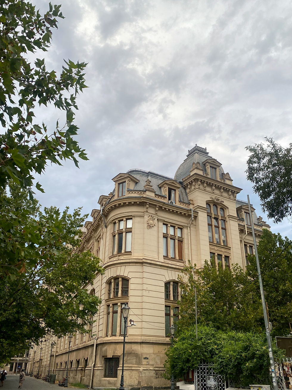 Historic beige building with arched windows, surrounded by green trees under a cloudy sky. Lamp posts and pathway in the foreground.