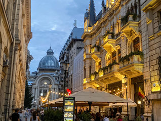Cobblestone street with ornate buildings at dusk, people strolling, diners under lit canopies, warm lights, and a clear, blue sky.