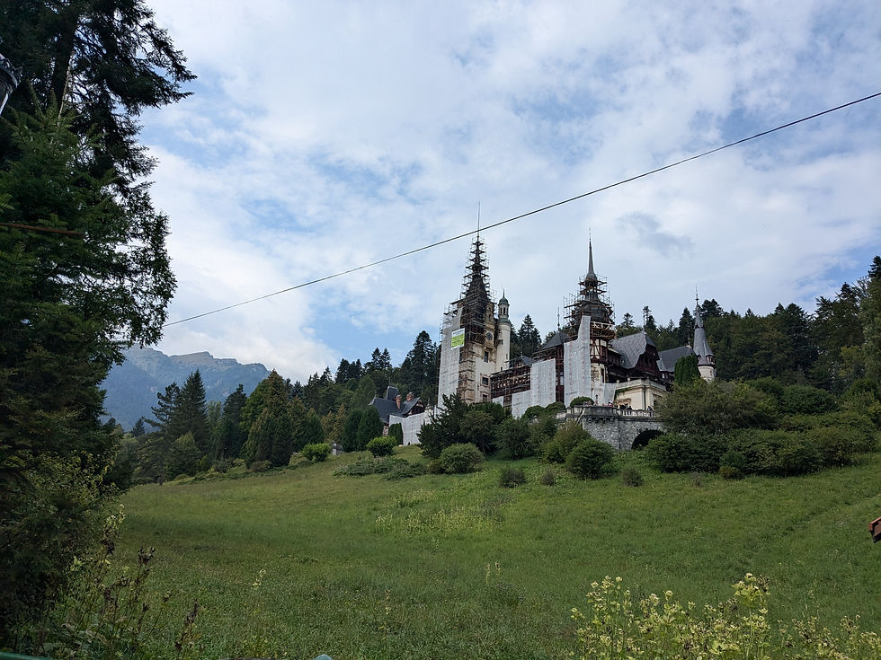 Peles Castle with towers under renovation stands on a lush green hill, surrounded by trees and mountains under a partly cloudy sky.
