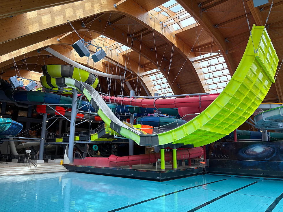 Indoor water park with colorful slides, including a neon green curve, above a clear blue pool. Wood ceiling and "Galaxy" mural visible.