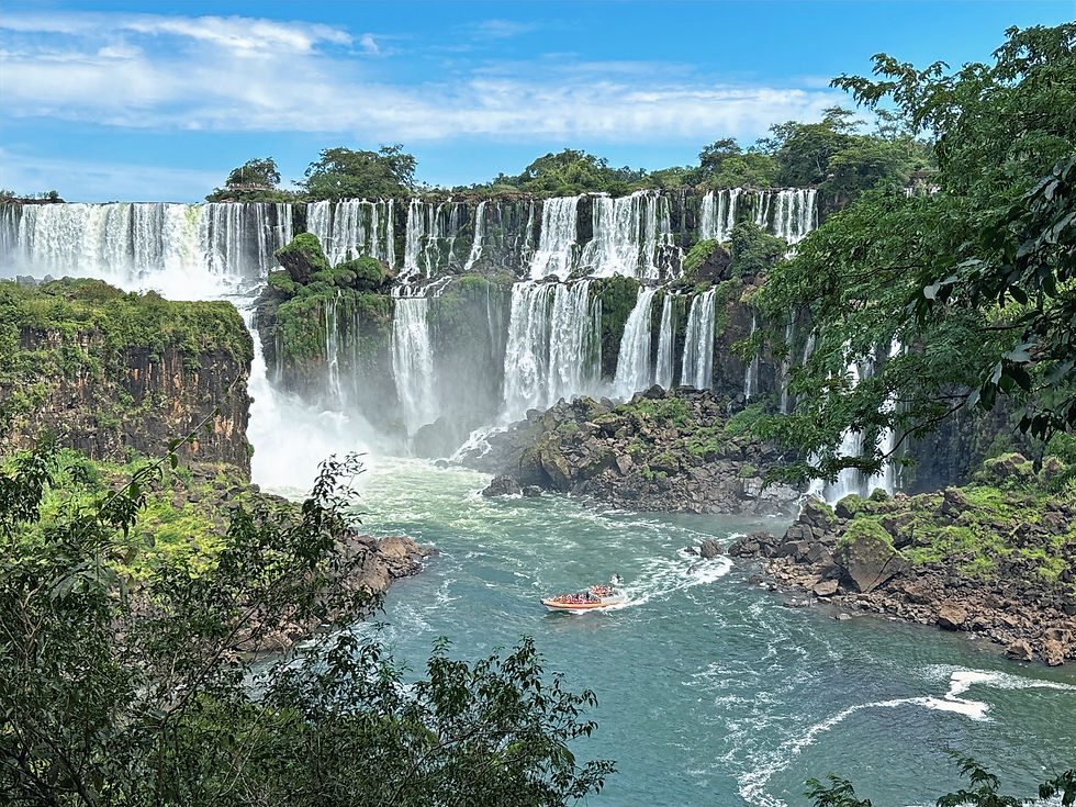 A boat on a river approaches a large, cascading waterfall surrounded by lush greenery and rocks under a blue sky.