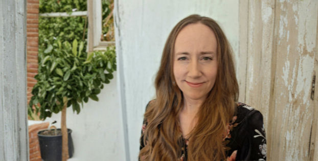 Woman (Shena) with long hair smiling softly indoors, wearing a floral top. Background includes a potted plant and rustic wooden walls.