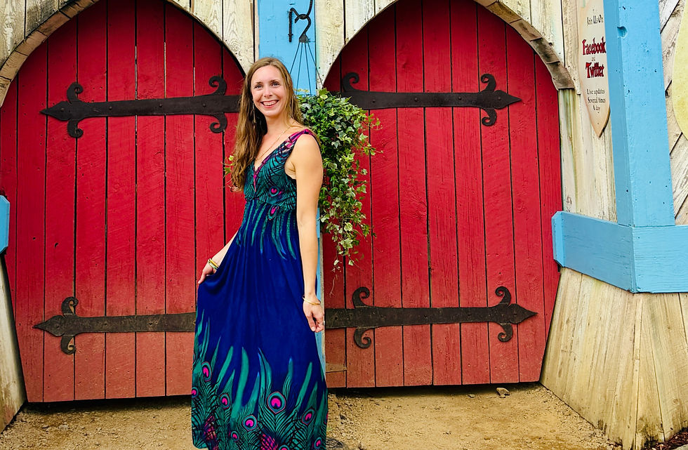 Woman (Sarah) in a blue dress with peacock patterns smiles in front of red wooden doors with black hinges. Hanging plants and rustic setting.