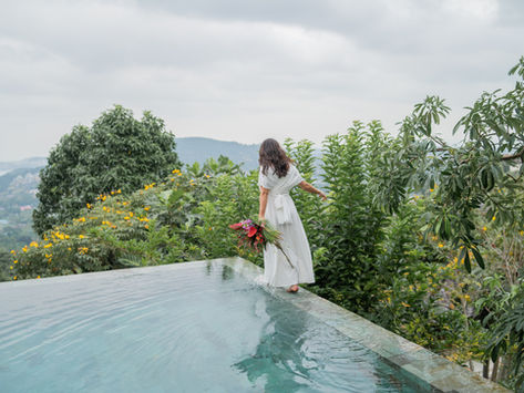 bride in a white dress balances on the edge of an infinity pool, holding tropical flowers. Lush greenery and thailand hills fill the serene background Panacea Retreat Villa in koh samui island in Thailand