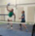 Two women play pickleball indoors. One in a green top hits a ball mid-air, while the other in blue watches. Both wear knee braces.