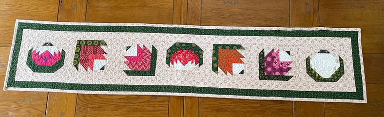 A handmade table runner featuring patchwork blocks made from Shweshwe fabrics. Each block highlights a different colour and pattern, bringing texture and heritage design to the table.