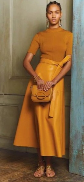 Modelo vestindo look monocromático cor caral=melo alaranjado. Ela veste rasteirinha dourada, saia midi de couro, blusa de manga 3/4 e uma bolsa de couro.
