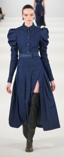 Modelo desfilando na passarela com um vestido midi de manga comprida jeans. O vestido tem uma fenda na lateral e a modelo também usa uma bota over the knee preta.