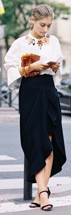 Mulher andando na rua e digitando no celular, vestindo saia midi preta e blusa branca com vários broches de flores marrom e laranjas e uma bolsa marrom.