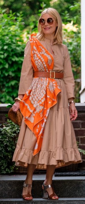 Mulher de meia idade loira vestindo vestido midi bege com cinto e sandália marrom e um lenço laranja.