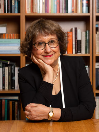 Portrait of a mature female journalist sitting at her desk, with a backdrop of books. This image conveys her experienced and knowledgeable demeanor, ideal for media profiles, editorial features, and personal branding. The books in the background emphasize her intellectual depth and commitment to journalism, showcasing her as a seasoned professional in her field