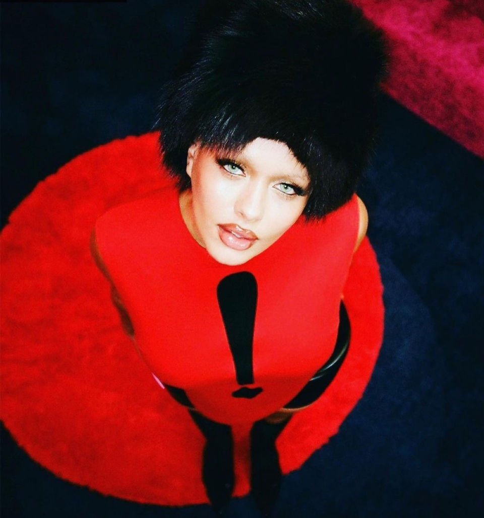 Woman wearing a red dress with an exclamation point with black fur hat, looking up.