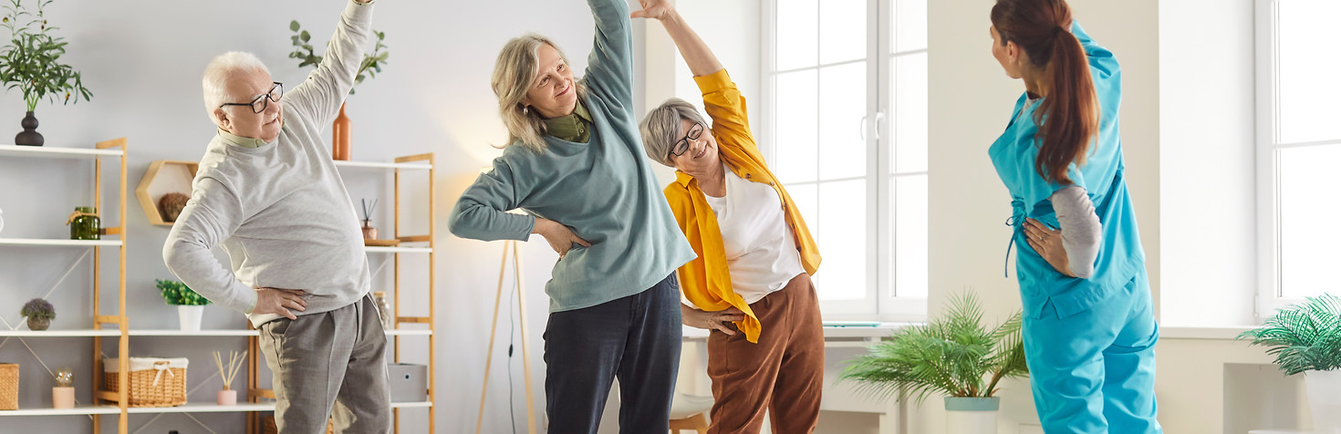 Groupe de personnes âgées faisant des exercices d’étirement guidés par une physiothérapeute dans une pièce lumineuse.