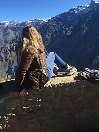 A woman with long hair sits on a stone wall, overlooking snow-capped mountains under a clear blue sky.