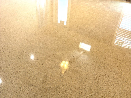 Polished beige floor reflects light in a room with exposed brick walls. A door and plastic-covered window in the background. Bright and clean.