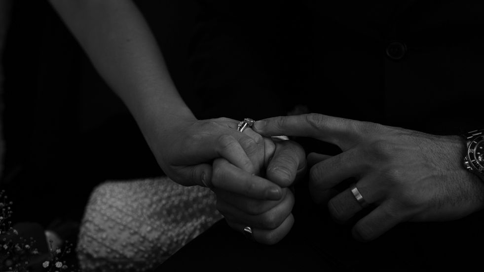 Black‑and‑white candid photo of the groom holding the bride’s hand in the taxi, gently touching her new wedding band on the way to the reception