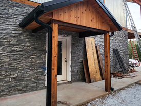 Marquesina de madera en la entrada, junto a revestimiento metálico y pared con aspecto de piedra, con materiales aún en el lugar.