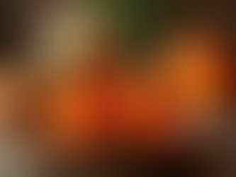 Orange bell peppers carved as jack-o'-lanterns, glowing on a wooden plate. More peppers and a plant in the blurred background. Halloween mood.