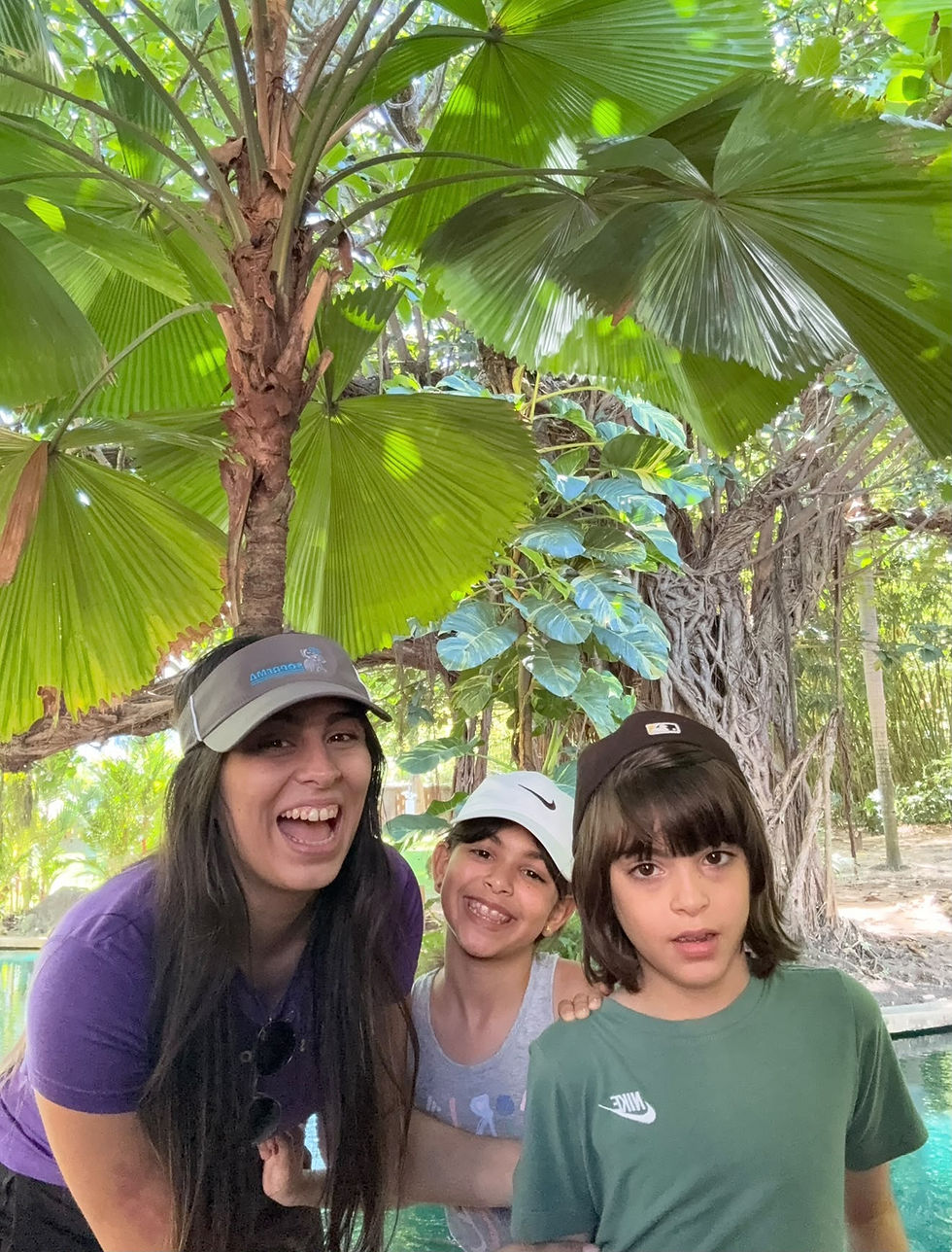 Luzmarie Pérez Torres posa con sus hijos Dylan y Mía al aire libre, rodeados de palmas y árboles.
