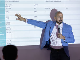 El Dr. José Caraballo Cueto señala datos estadísticos proyectados durante la presentación del estudio Movilidad Social en Puerto Rico, celebrada en la Fundación Luis Muñoz Marín el 12 de mayo de 2026. La pantalla muestra una tabla con distribución por clase social y grupos de edad.