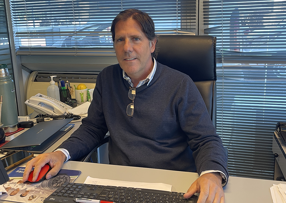 Hombre en oficina, sentado ante un escritorio con teclado y ratón rojo, mirando a cámara; persianas al fondo.