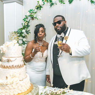 Bride and groom toast with champagne beside a white-and-gold wedding cake under a floral arch, with Mr & Mrs McCain sign.