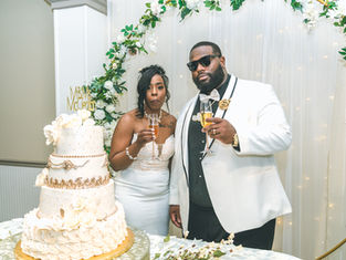 Bride and groom toast with champagne beside a white-and-gold wedding cake under a floral arch, with Mr & Mrs McCain sign.