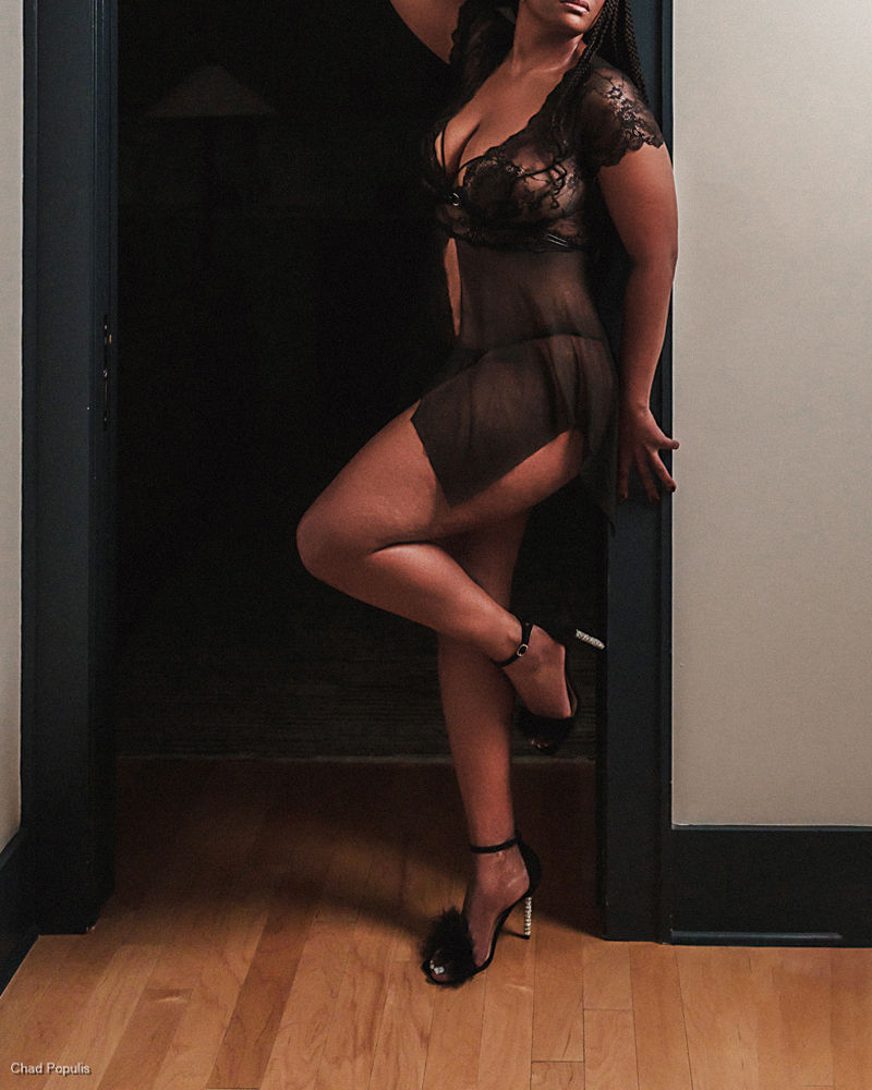 Woman poses in sheer black lingerie and heels in a doorway, with a dark room behind her and hardwood floor; Chad Populis watermark
