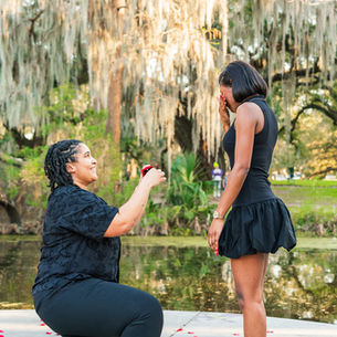 A person on one knee proposes with a ring in a park. Another stands, surprised, near a pond and bridge. Trees with moss in the background.
