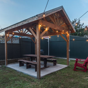 Wooden gazebo with string lights, picnic table, and red chair on grass. Surrounded by fence, trees, and house. Evening setting.