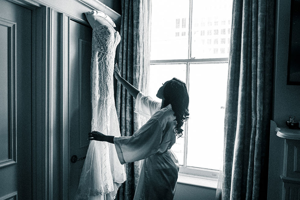 A woman in a robe admires a lace wedding dress hanging by a bright window. The room has elegant paneling and floral curtains, creating a serene mood.