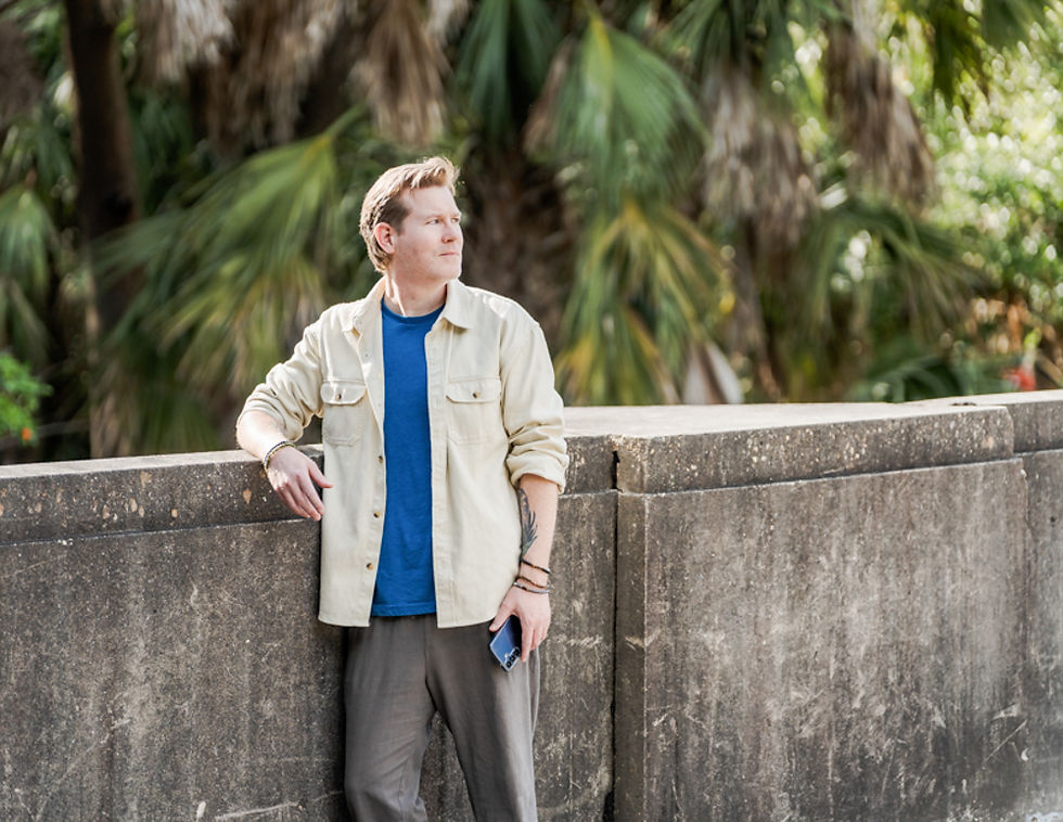 Man in a beige jacket leans on a stone wall, holding a phone, with palm trees in the background. He appears thoughtful, wearing a blue shirt.
