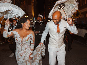 Wedding Couple second line parades down a New Orleans street at night. They hold ornate umbrellas, smiling, with a brass band playing behind them.