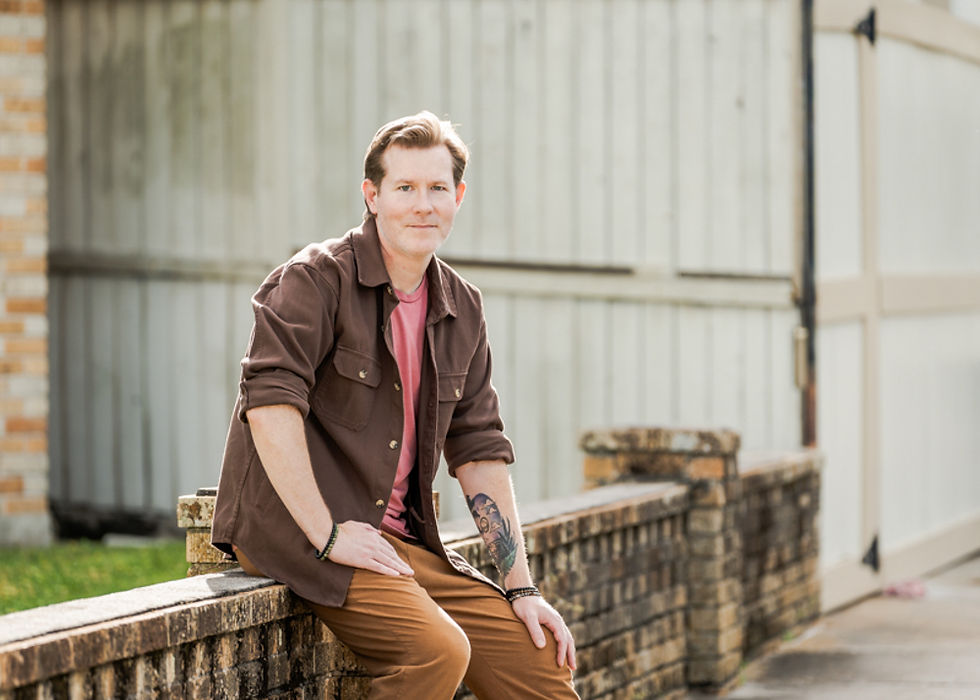 Man sitting on a brick wall wearing a brown jacket. Background features a wooden fence and brick house. Relaxed, casual mood.