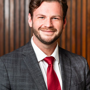 Man smiling in a gray suit and red tie, standing against a wooden panel background. The mood is professional and friendly.