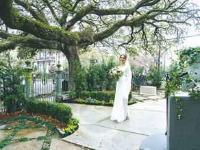 Bride in white dress holding bouquet under large tree, surrounded by lush garden, with elegant iron gate in the background. Peaceful mood.