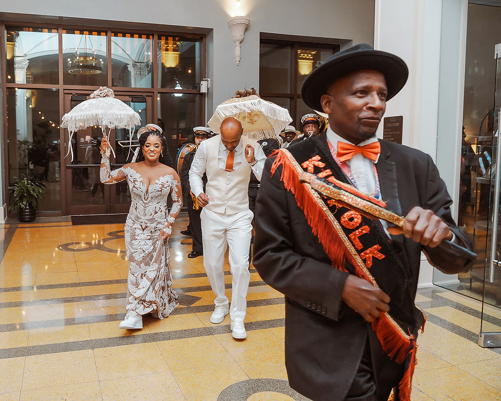 Bride in lace gown and groom in white suit enter a grand lobby under umbrellas, led by a man in black hat and orange sash.