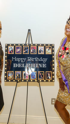 Two smiling women pose beside a lit Happy Birthday Delphine sign at a festive party, one in black and one in gold sequins.