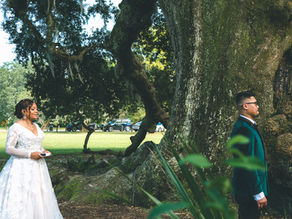 Bride in white gown stands behind groom in green jacket for a First look near large tree, grassy Audubon park setting with cars in background. Romantic mood at the Tree of Life.