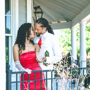 A couple embraces on a porch at Kenner City Pavillion. The woman wears a red dress and gloves; the man wears a white shirt and tie. Columns and greenery surround them.