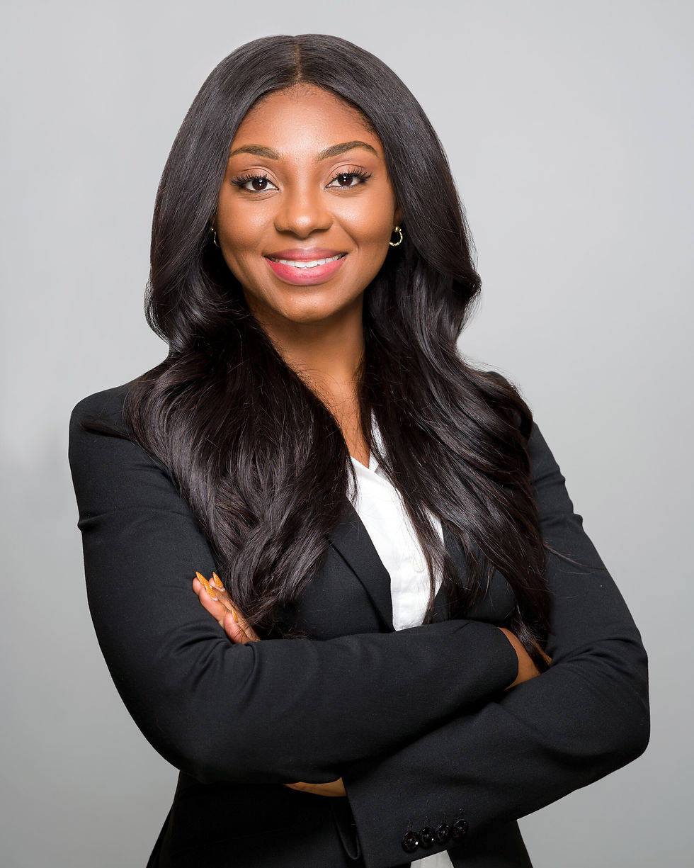 Woman in black blazer and white shirt smiles confidently with arms crossed. Plain gray background, long dark hair, professional setting.
