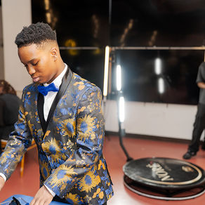 Young man in a blue-and-gold floral suit and bow tie adjusts items at a table in an event space, with a lit backdrop behind him.