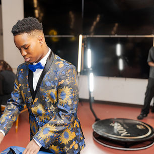 Young man in a blue-and-gold floral suit and bow tie adjusts items at a table in an event space, with a lit backdrop behind him.