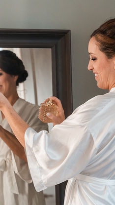 Bride in white robe smiles in mirror while applying perfume. "BRIDE" text on robe. Soft light and calm setting.