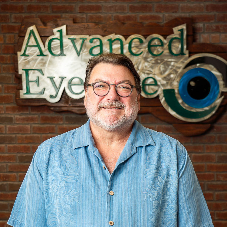 Smiling man in glasses and a blue shirt stands before an Advanced EyeCare sign on a brick wall.