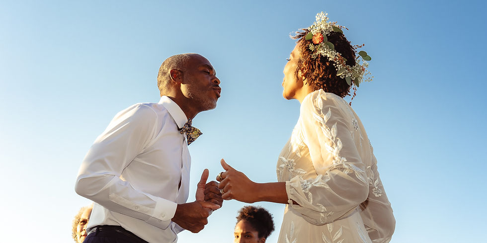 A couple dances under a clear blue sky. The woman wears a floral crown and cream dress, and the man wears a white shirt and bow tie. Joyful mood.