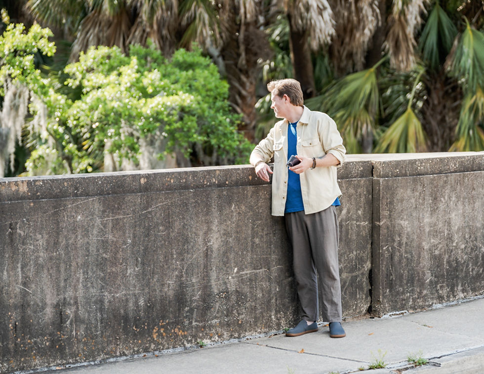 Man in casual attire leans on a concrete wall, holding a phone, looking left. Green foliage in the background, bright and sunny.