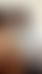 A smiling couple sits on a bed. She wears a tan sweater, he a white shirt. Warm light fills the room, creating a cozy atmosphere.