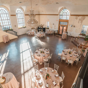 Treme Market Branch. Elegant event hall with round tables, pink tablecloths, and white chairs. Large windows, chandelier, and natural light create a warm ambiance.